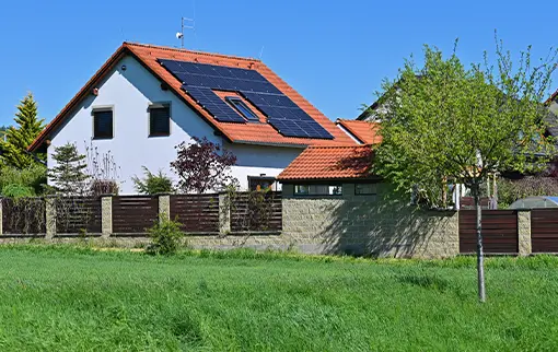 Dom jednorodzinny z panelami solarnymi zamontowanymi na czerwonej dachówce, widoczny za ogrodzeniem i zieloną łąką