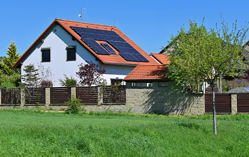 Nowoczesne panele fotowoltaiczne Solemark zamontowane na czerwonym dachu wielorodzinnego domu mieszkalnego, widocznego za płotem i zieloną łąką